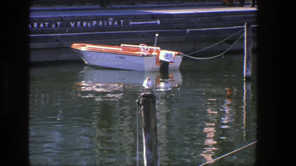 1975 Denmark 8mm frame with a seagull on a wooden harbor post and a small motorboat tied along the quay behind it.