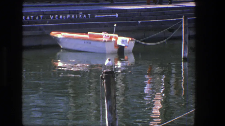 1975 Denmark harbor frame showing a moored white and red motorboat, a seagull on a post, and shimmering reflections on the water.