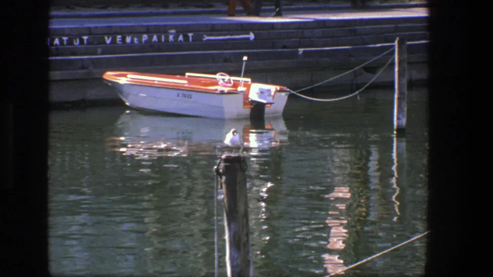 1975 Denmark waterfront frame with the small motorboat, taut mooring line, quay sign, shimmering water, and gull in the foreground.