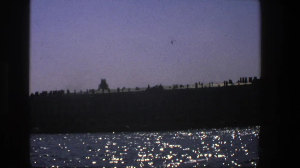 1975 Denmark 8mm frame of a dark waterfront building line, pale sky, and scattered sunlight on the water.