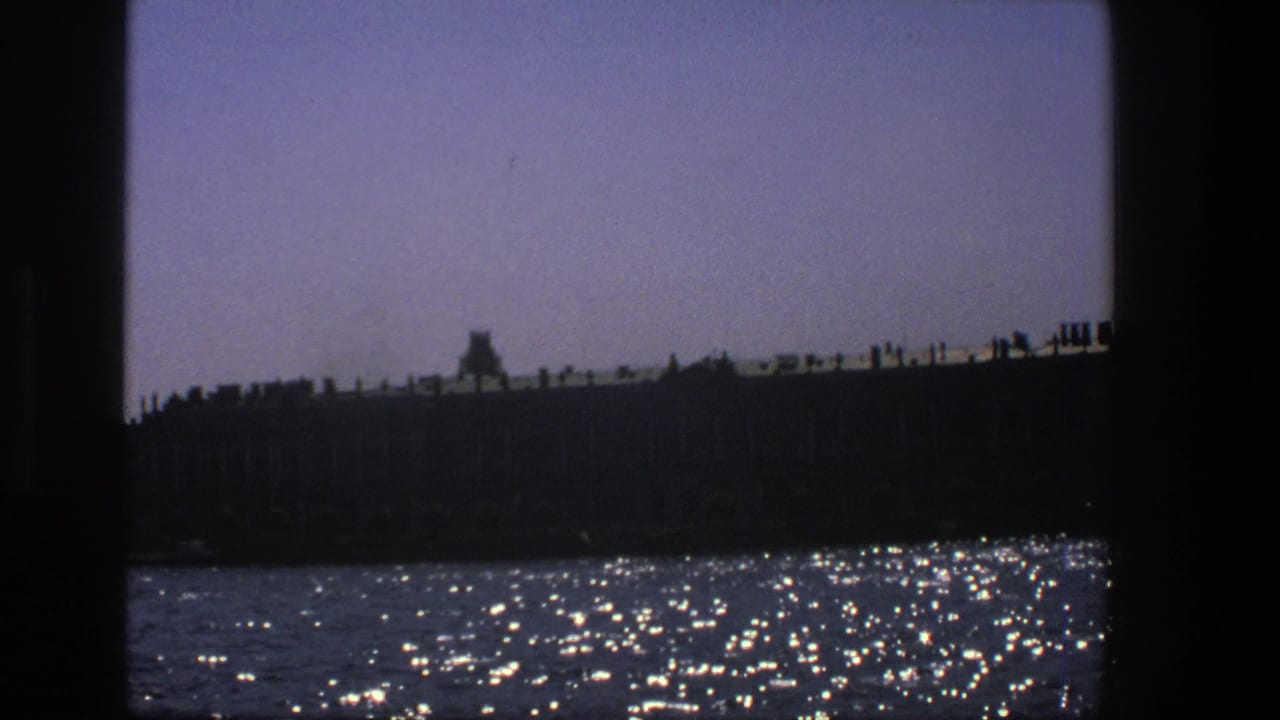 1975 Denmark 8mm frame showing a shadowed waterfront seen from water, with bright reflections and dark vertical borders.