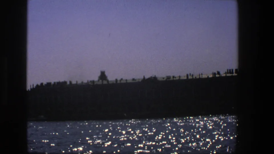 1975 Denmark 8mm frame showing a shadowed waterfront seen from water, with bright reflections and dark vertical borders.