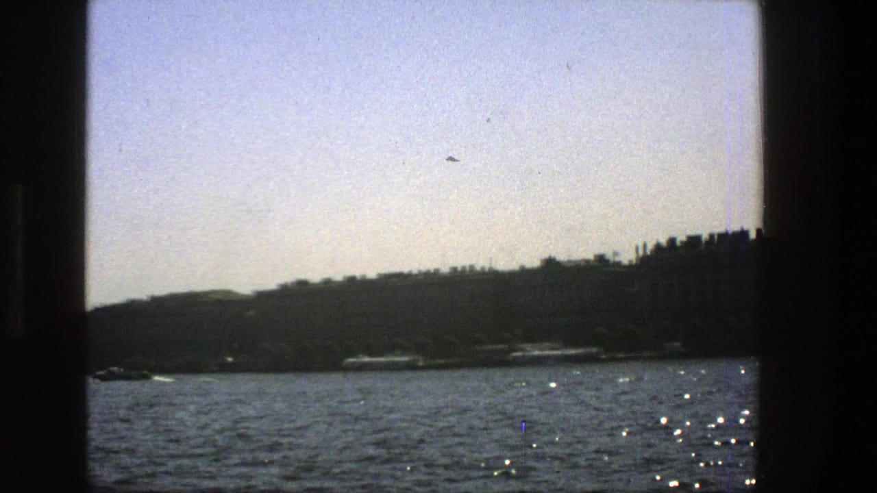 1975 Denmark home-movie view across sunlit water toward a dark shoreline, seen through the edge of a vessel window.