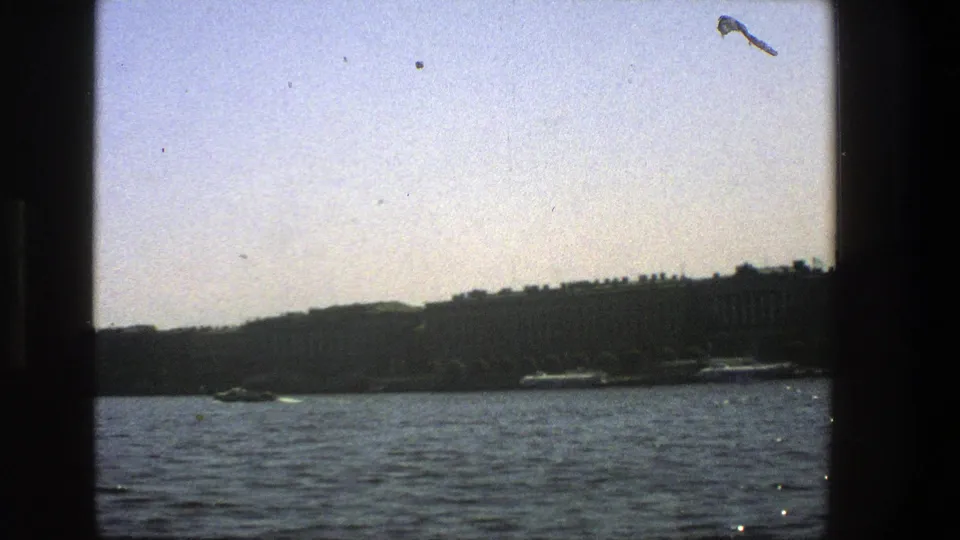 Vintage 8mm view from a vessel in Denmark, looking across rippled water toward a low dark shoreline and moored or passing boats.