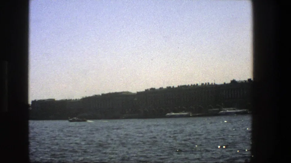 1975 Denmark home-movie frame of shimmering water, distant shoreline buildings, and small boats, partially bordered by dark vessel framing.