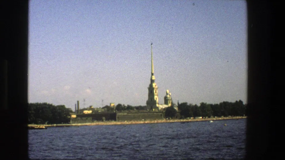 1970s 8mm frame of a historic-looking waterfront with a tall spire, trees and blue water in the foreground.