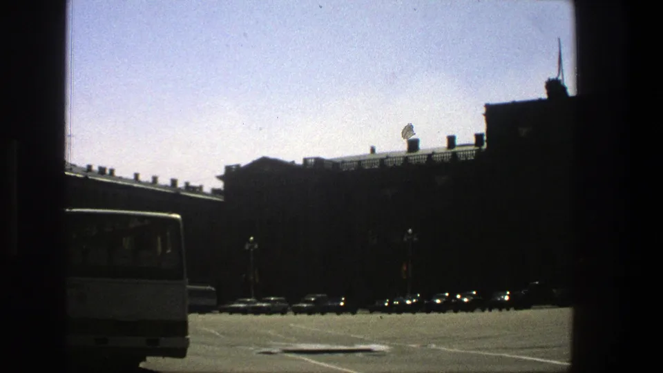 1975 8mm frame shifts to a city street with a white bus, parked cars, dark building silhouettes, and a pale blue sky.