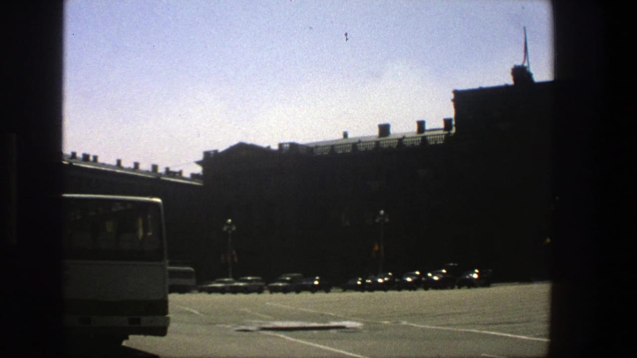 1975 Denmark 8mm frame showing a broad sunny parking area, a white bus edge, parked cars, and a large dark building.