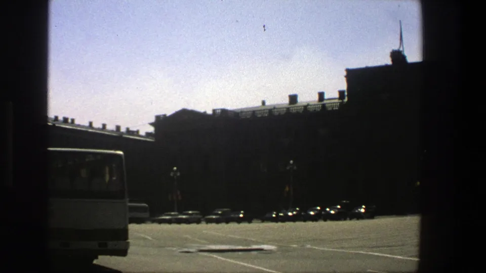 1975 Denmark 8mm frame showing a broad sunny parking area, a white bus edge, parked cars, and a large dark building.