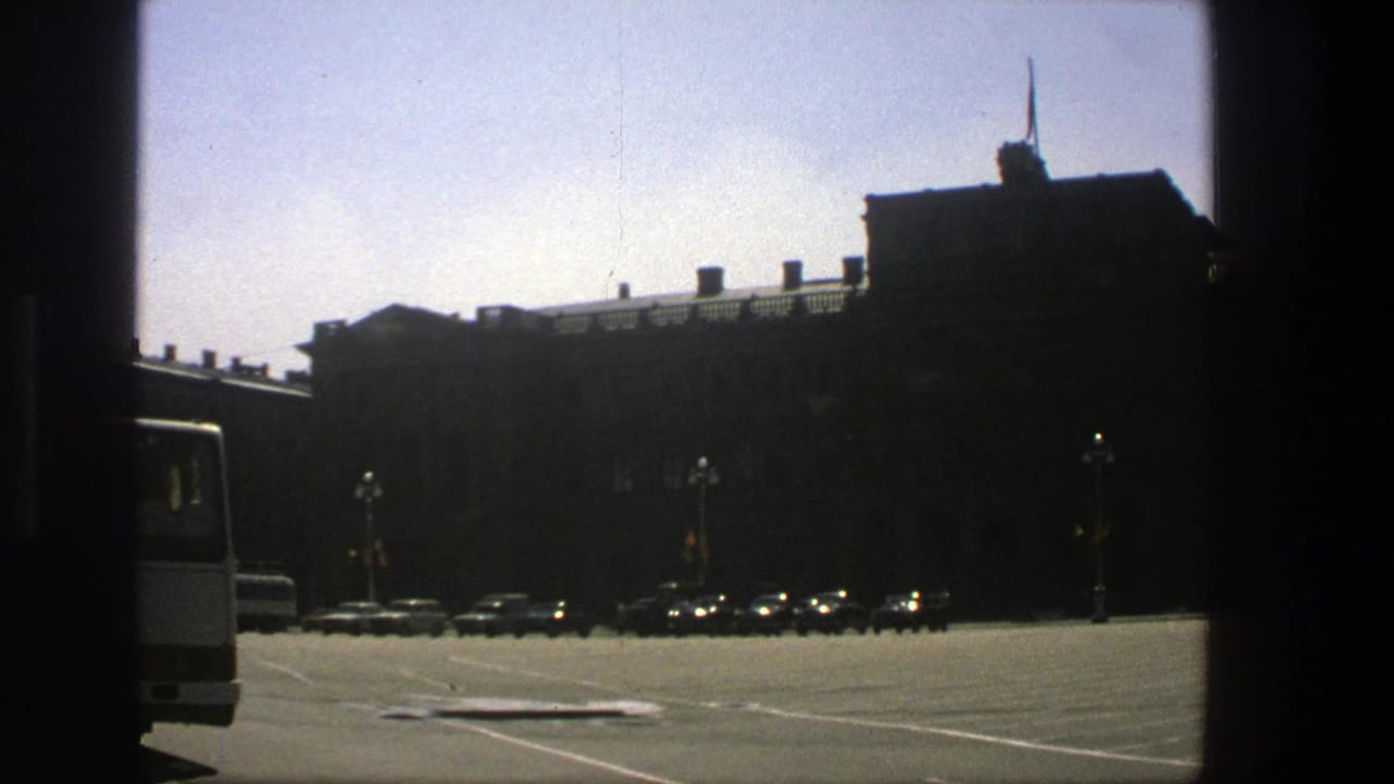 Wide paved area with parked cars, lampposts, and a dark building in bright sun.