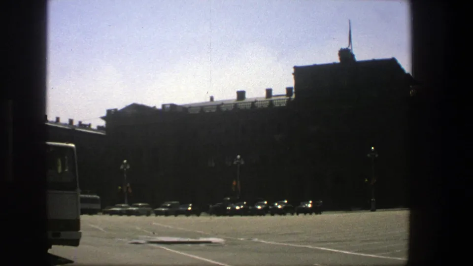 1975 Denmark frame with a sunlit open parking area, vintage cars lined near the curb, lampposts, and a shadowed building facade.