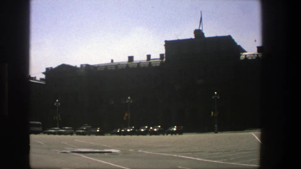 1975 Denmark 8mm view of a large shadowed building, a row of parked cars, lampposts, and strong daylight across pavement.