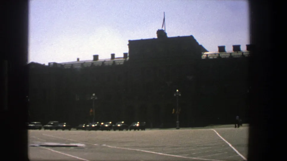 1975 Denmark frame showing parked cars before a dark building, bright pavement, lampposts, and two very small distant people at right.