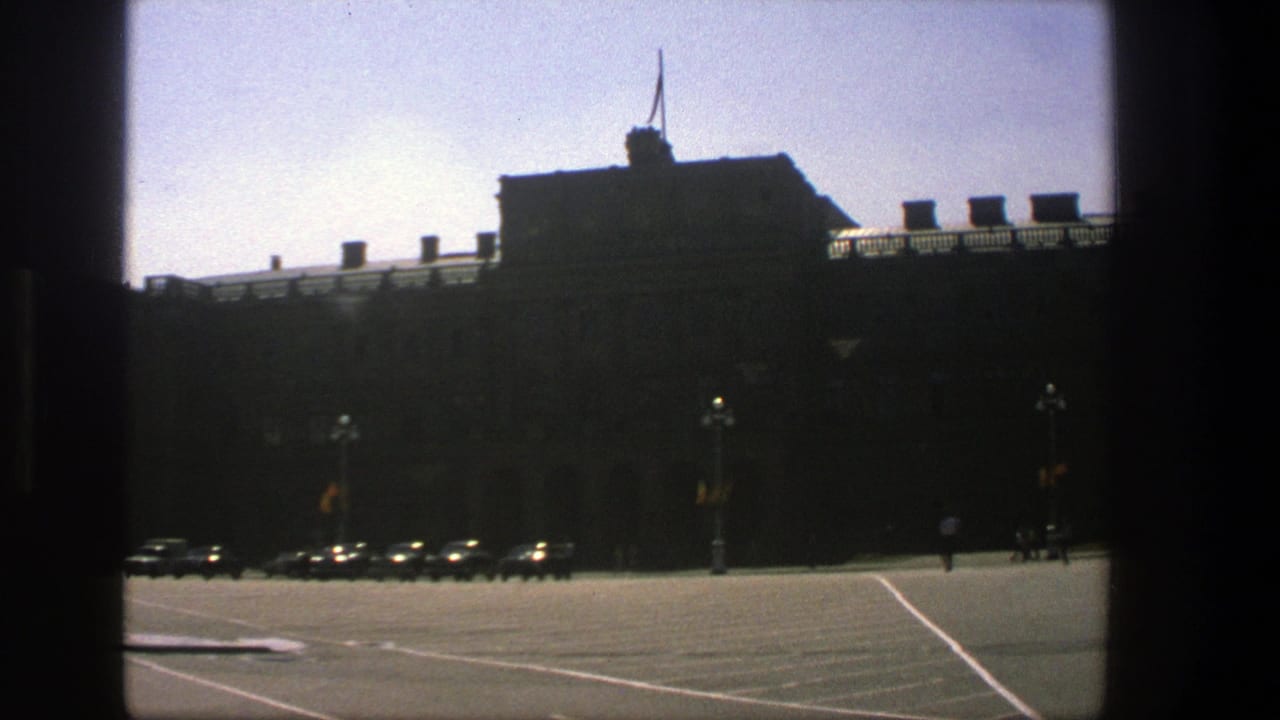 1975 Denmark 8mm view of a large silhouetted building beside a broad street with parked cars and small pedestrian figures.