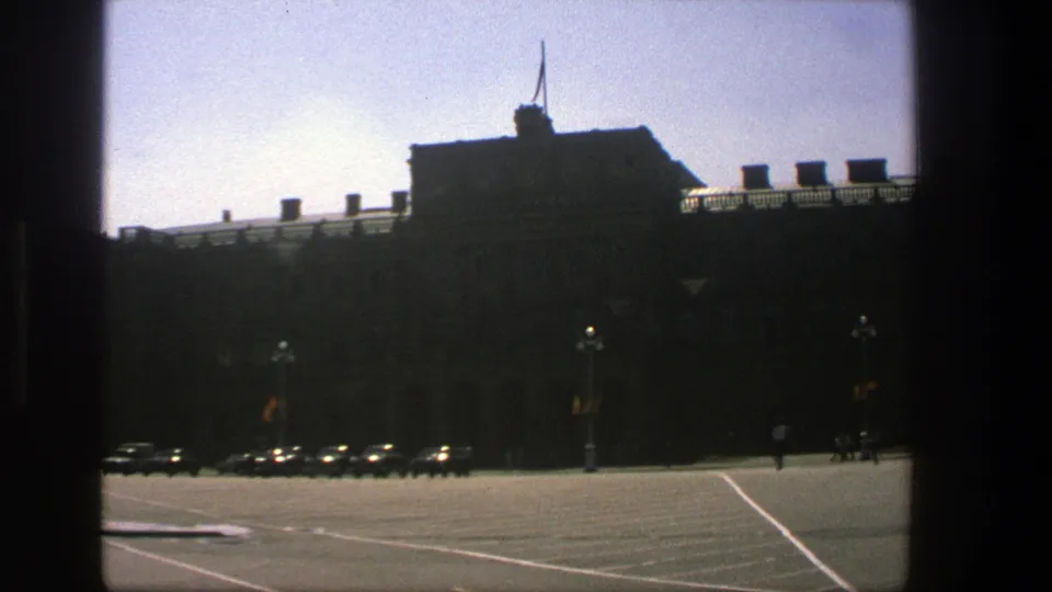 1975 Denmark 8mm view of a large silhouetted building beside a broad street with parked cars and small pedestrian figures.