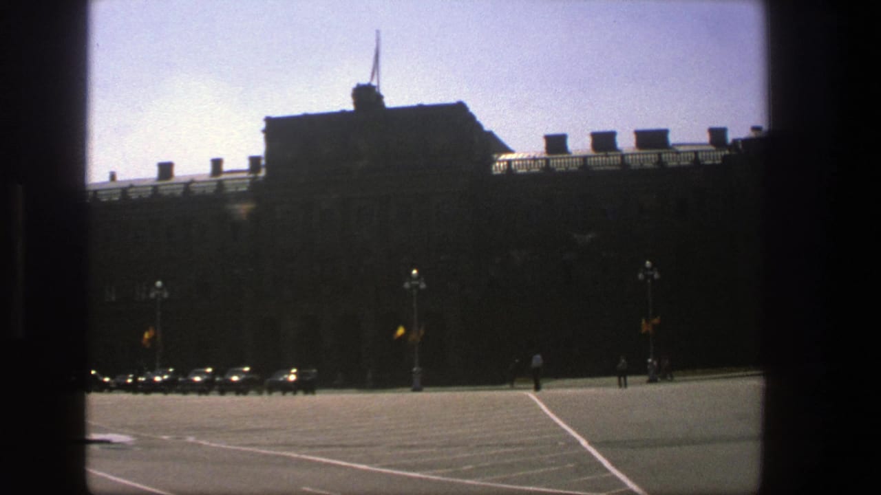 Wide street view with the large building in shadow, parked cars, lamp posts, and pedestrians.