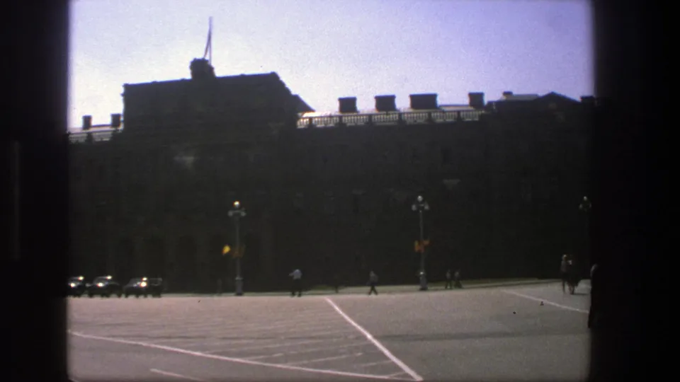 In this 1975 Denmark frame, a broad street and tall lamps sit before a dark building facade with small pedestrians crossing the scene.