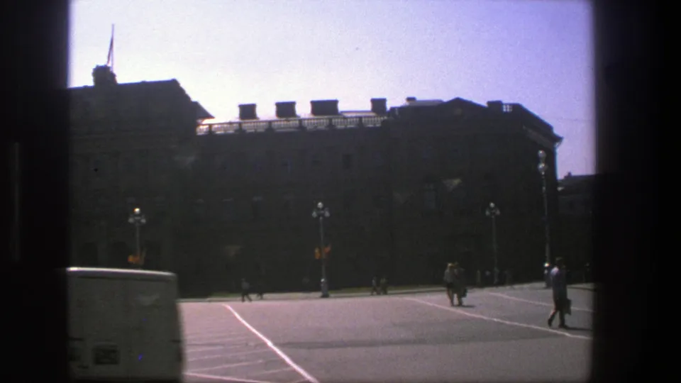 A white van enters the foreground of the 1975 Denmark street view, with the large building and distant pedestrians behind it.