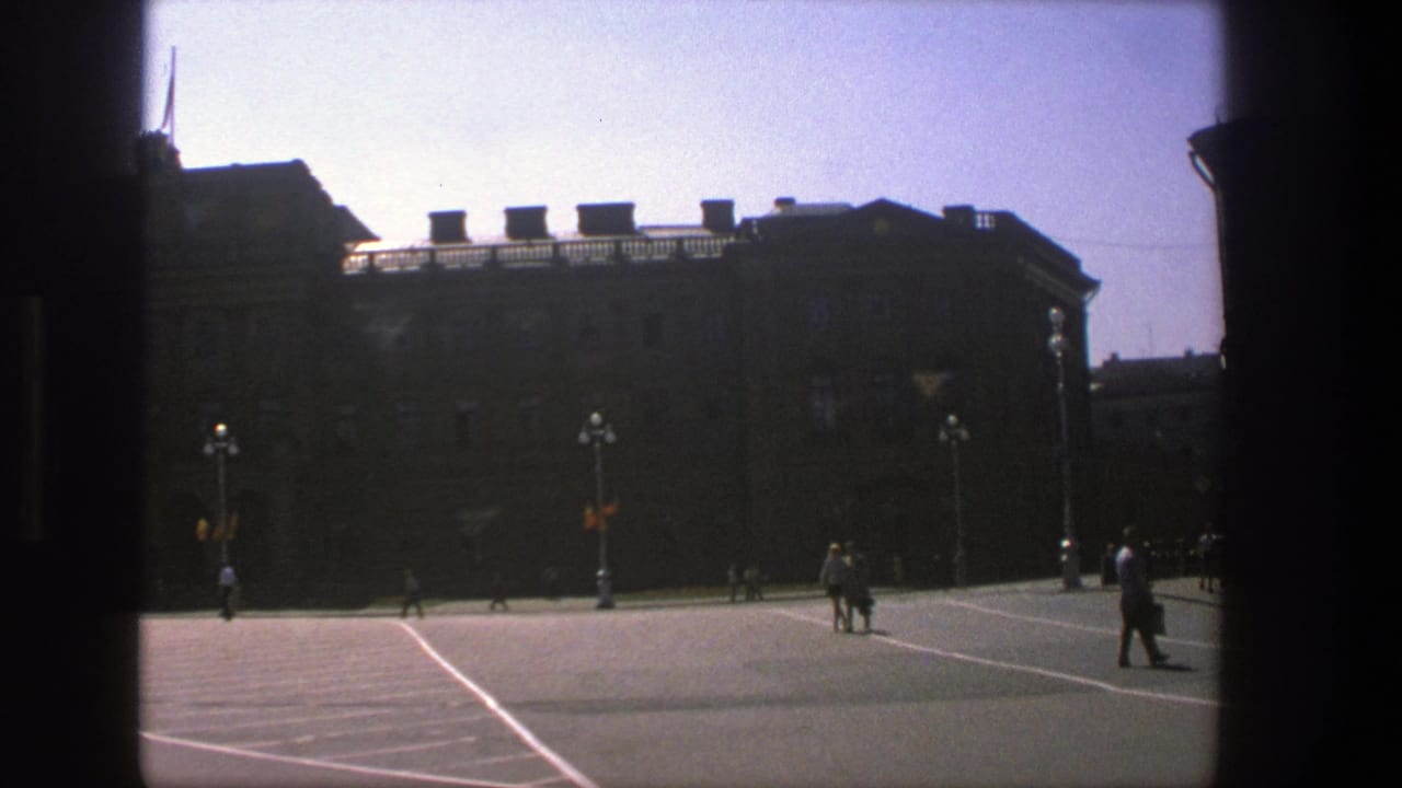 1975 Denmark home-movie frame showing a sunlit public square, distant pedestrians, ornate lamps, and a shadowed historic building.