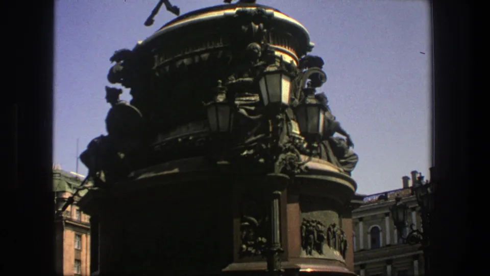 The 8mm frame shows the monument base in shadow, ornate lamps, rooftop lines, and a clear sky in a visitor-shot composition.