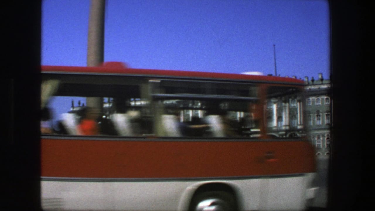Red-and-white bus passes in front of an ornate civic building under blue sky.