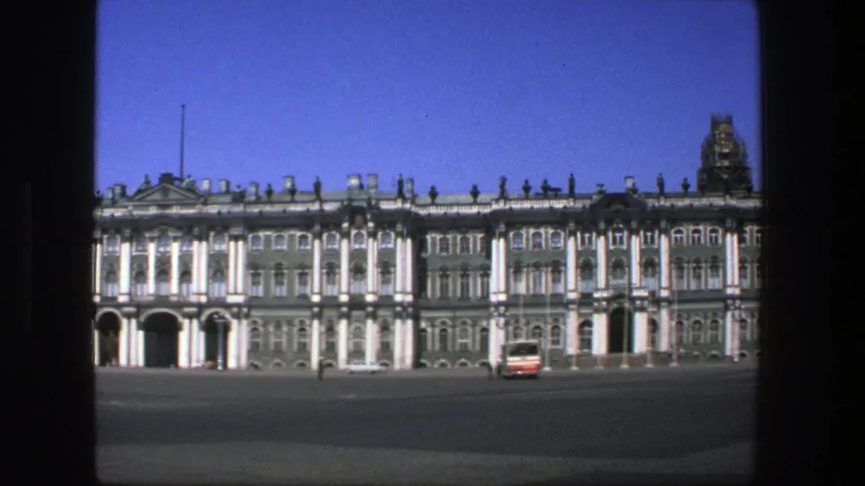 1975 8mm travel frame showing the long historic building facade, a red-and-white bus, and small distant pedestrians in the square.