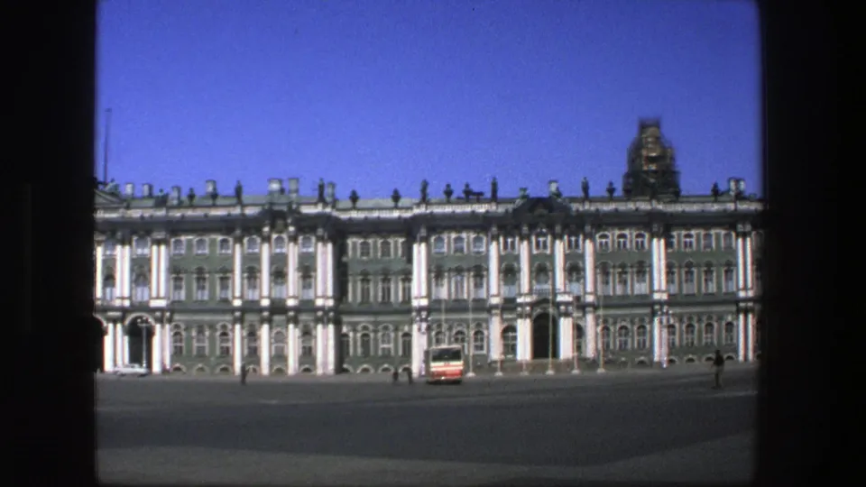 1975 8mm frame of a broad historic facade and open plaza, with a red-and-white bus and distant people visible.