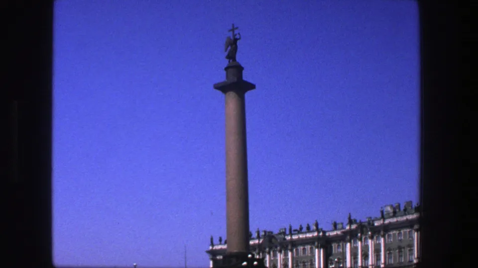 A tall monument column with a statue is framed against blue sky, with part of the palace visible below.