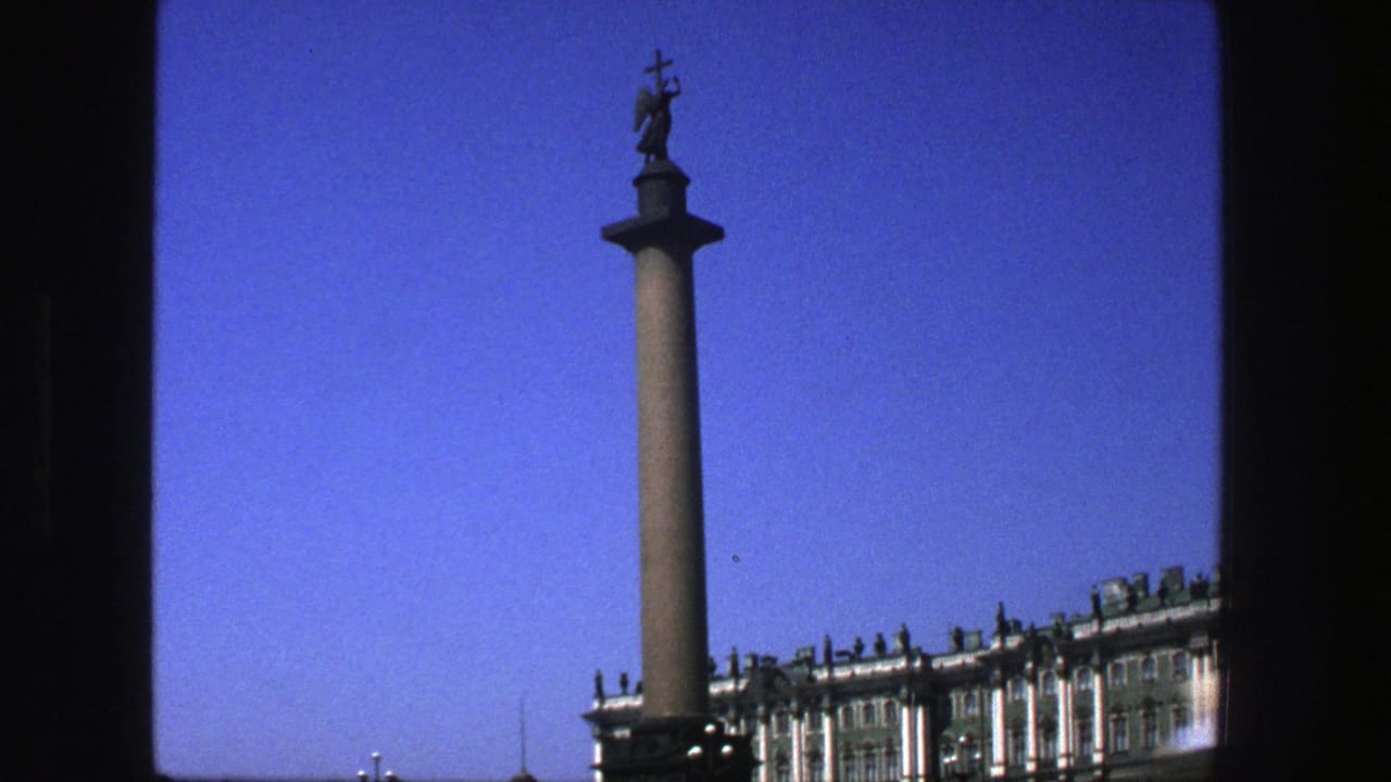 1975 8mm travel frame showing an angel-topped column and formal architecture against a deep blue sky, with dark film borders visible.