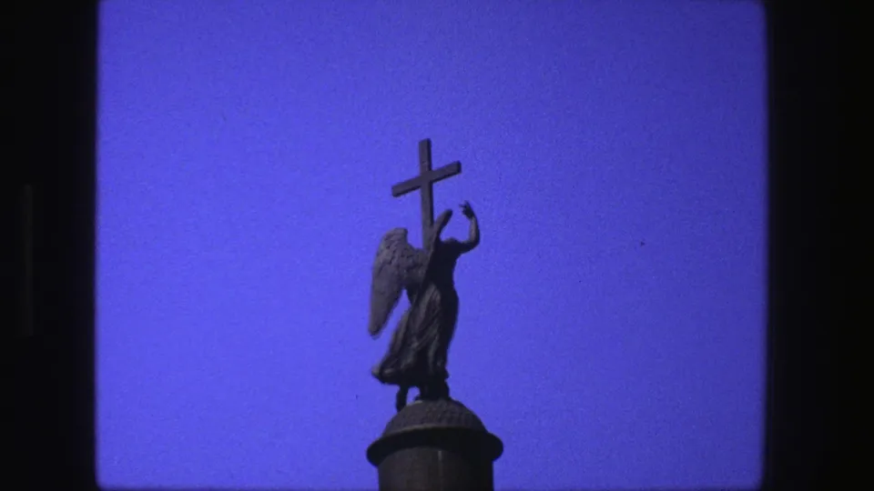 Close 1975 8mm frame of the angel figure and cross silhouetted against blue sky, with the column top partly visible.