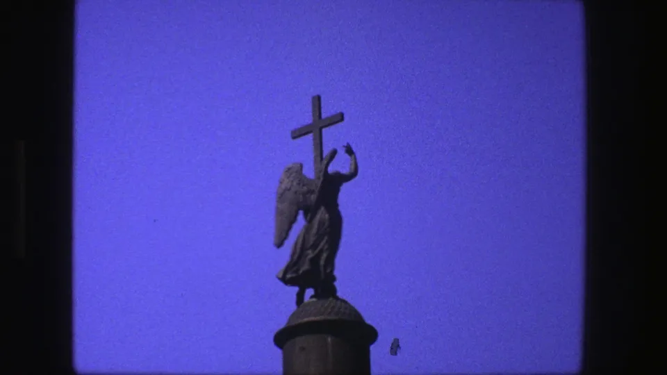1975 8mm close view of the winged statue holding a cross, with saturated blue sky and slight film speckling.