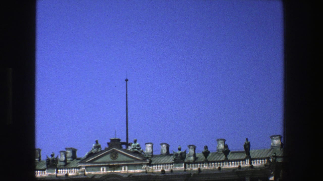 Historic roofline with statues, chimneys, pediment, and flagpole against a blue sky.