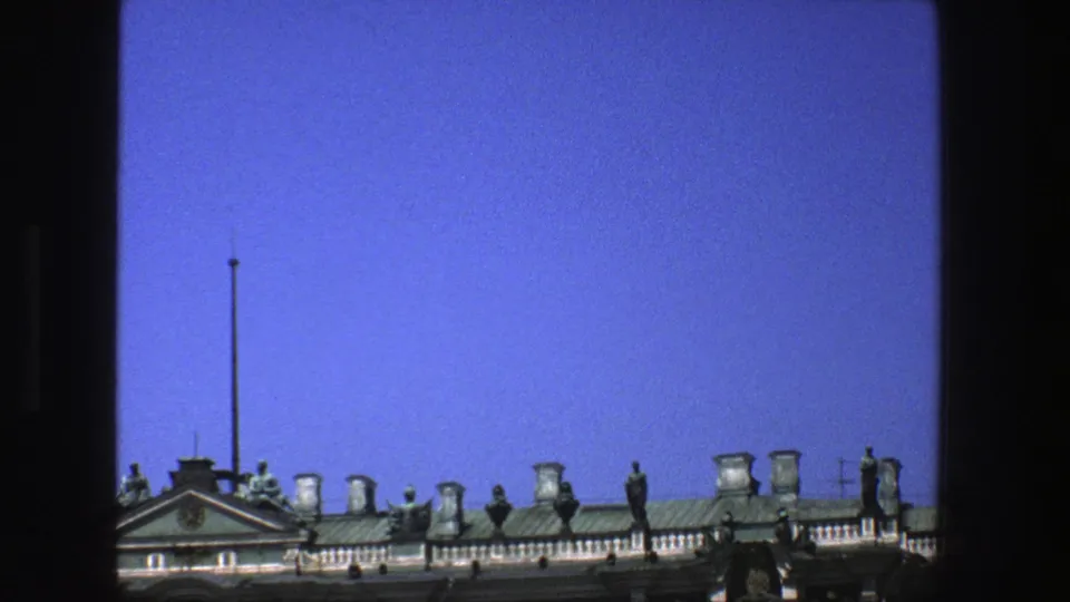 1975 Denmark 8mm frame with the roofline set low in view, showing a pediment, tall flagpole, statues, chimneys, and open blue sky.