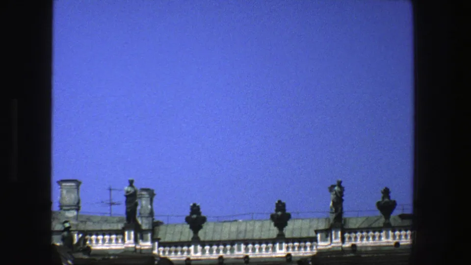 1975 Denmark home-movie view of a historic rooftop balustrade with statues, urn-like ornaments, chimneys, and saturated blue sky.