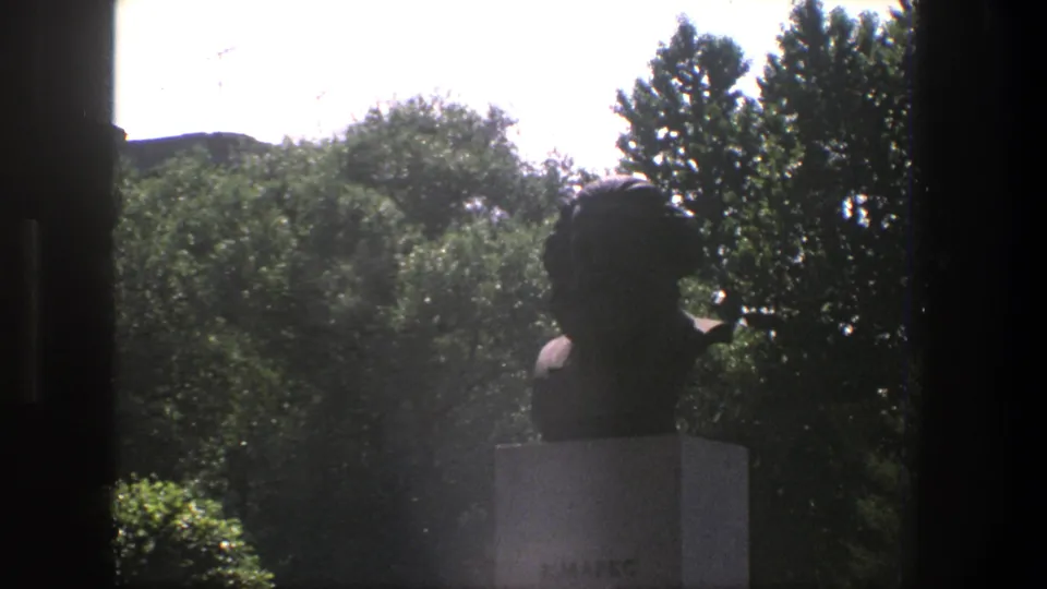 1975 Denmark 8mm frame showing a silhouetted bust monument on a pale pedestal, partly obscured by dark window framing and backed by trees.