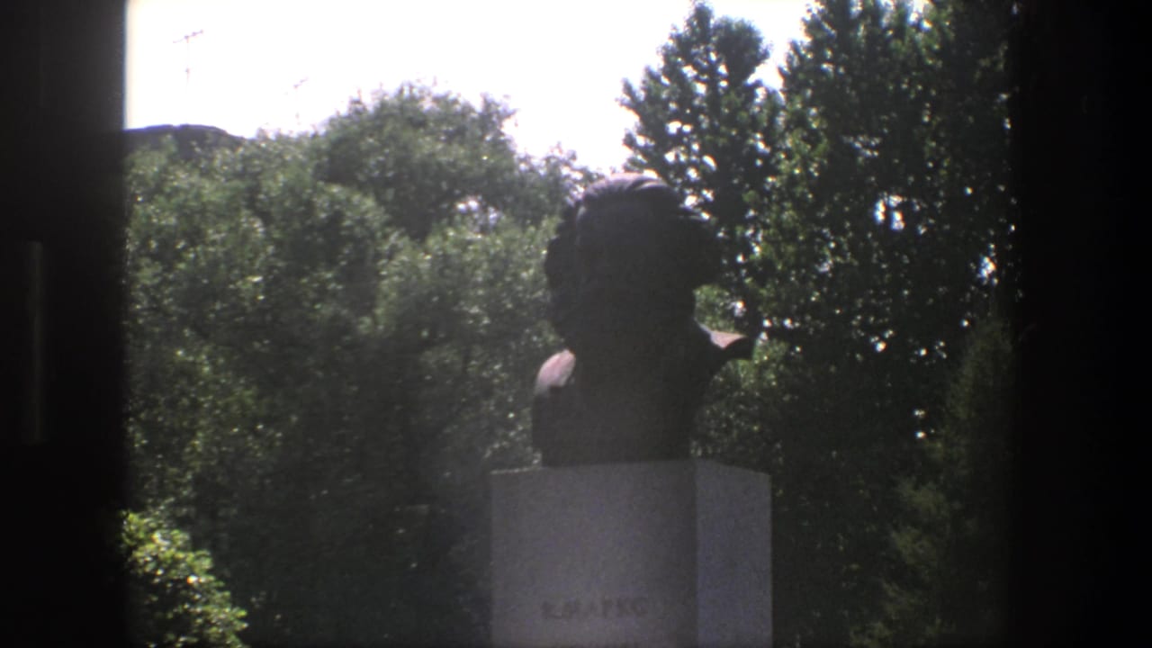 1975 Denmark frame with a dark bust on a stone pedestal, seen through a window-like opening against dense green trees and bright sky.