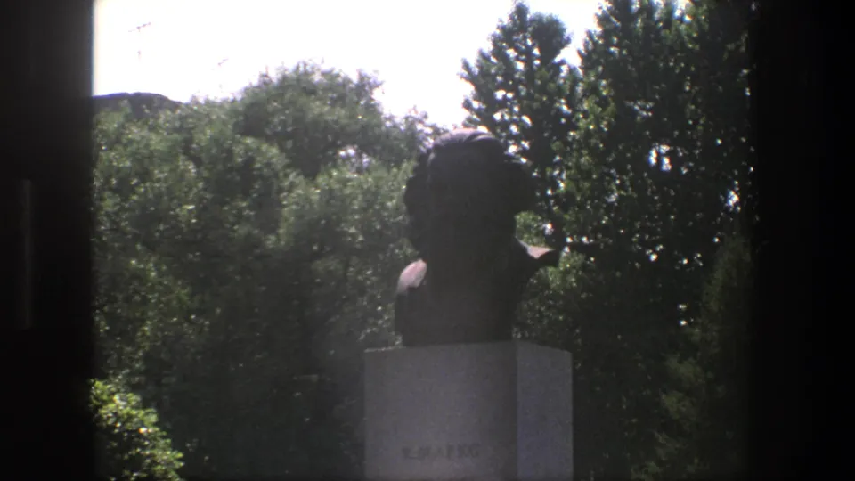 1975 Denmark frame with a dark bust on a stone pedestal, seen through a window-like opening against dense green trees and bright sky.