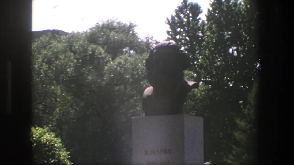 1975 Denmark 8mm view of the bust monument from a visitor vantage point, with the pedestal base, trees, and dark window framing visible.