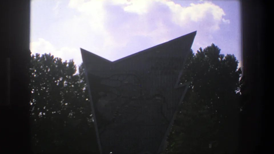 1975 Denmark frame showing an angular engraved stone marker beside trees, with dark framing at the edge and bright sky above.
