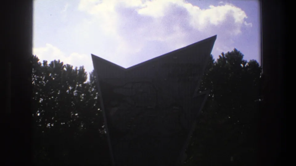 1975 Denmark 8mm frame showing a tall angular landmark in silhouette against a bright clouded sky and tree line.