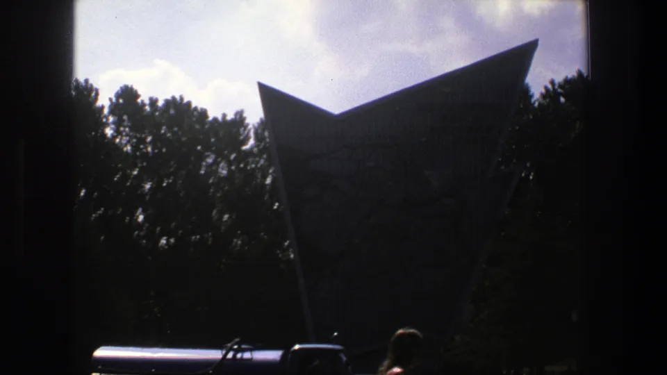 A blue tanker truck appears at the base of the angular Denmark landmark, with trees and bright sky behind it.
