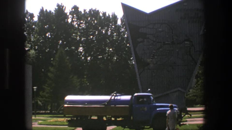 A blue diesel tanker truck is parked in front of the tall angular Denmark landmark, with one person visible near the right side.