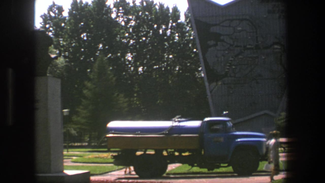 Blue tanker truck parked near a tall monument and trees.