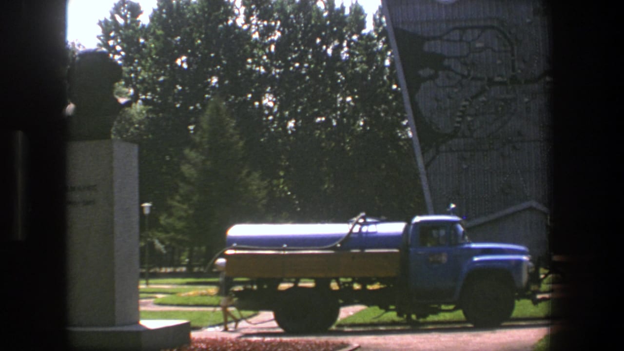 Blue tanker truck parked beside a monument in a landscaped area.