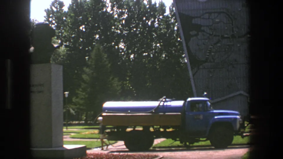 Blue tanker truck and tall monument seen in a 1975 Denmark home-movie frame, with trees and lawn behind.