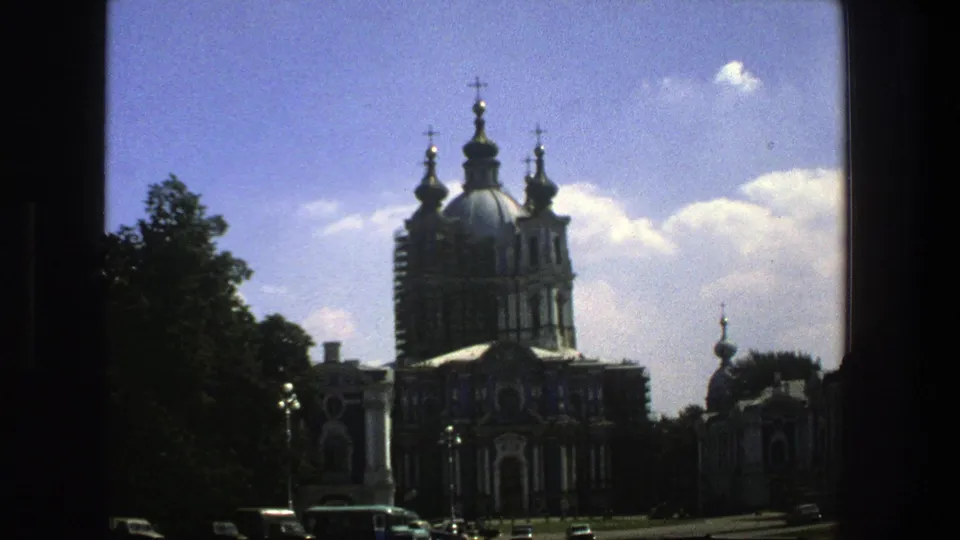 1975 8mm travel frame in Denmark showing the ornate church from farther right, with domes, crosses, trees, cars, and bright cloud cover.