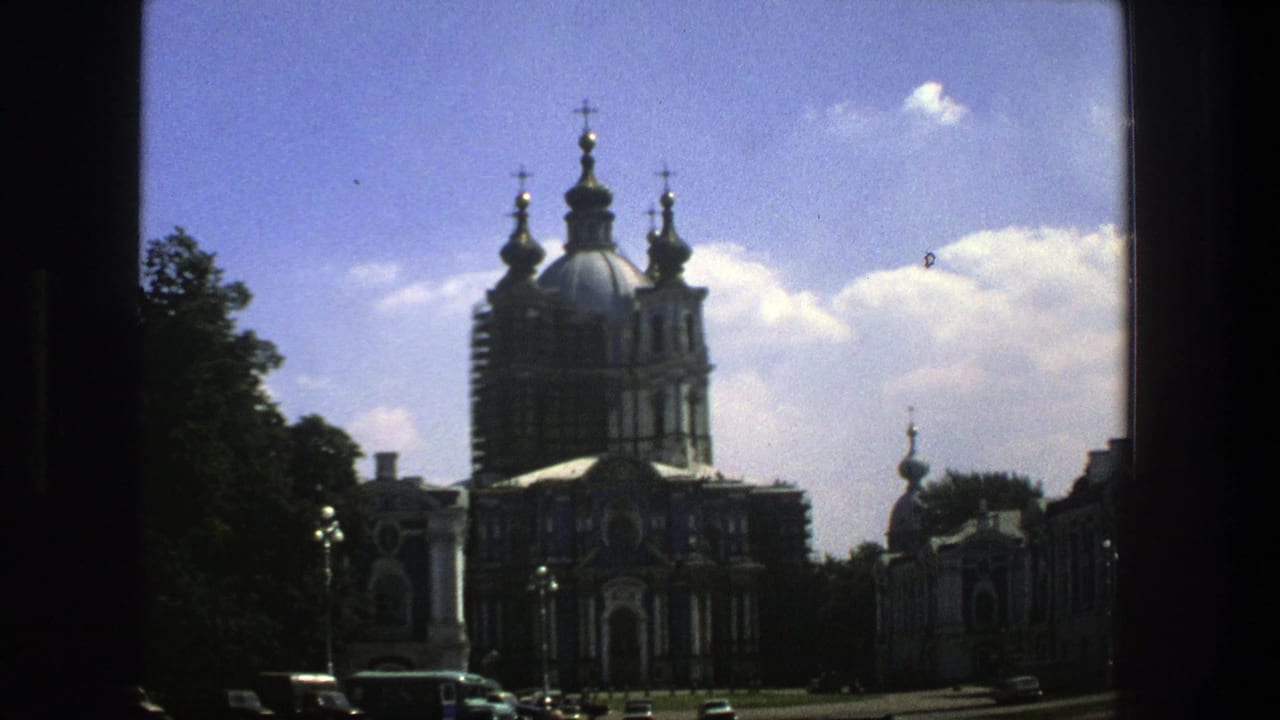 1975 8mm view in Denmark showing a large Orthodox church with blue onion domes, gold crosses, trees, parked cars, and blue sky.