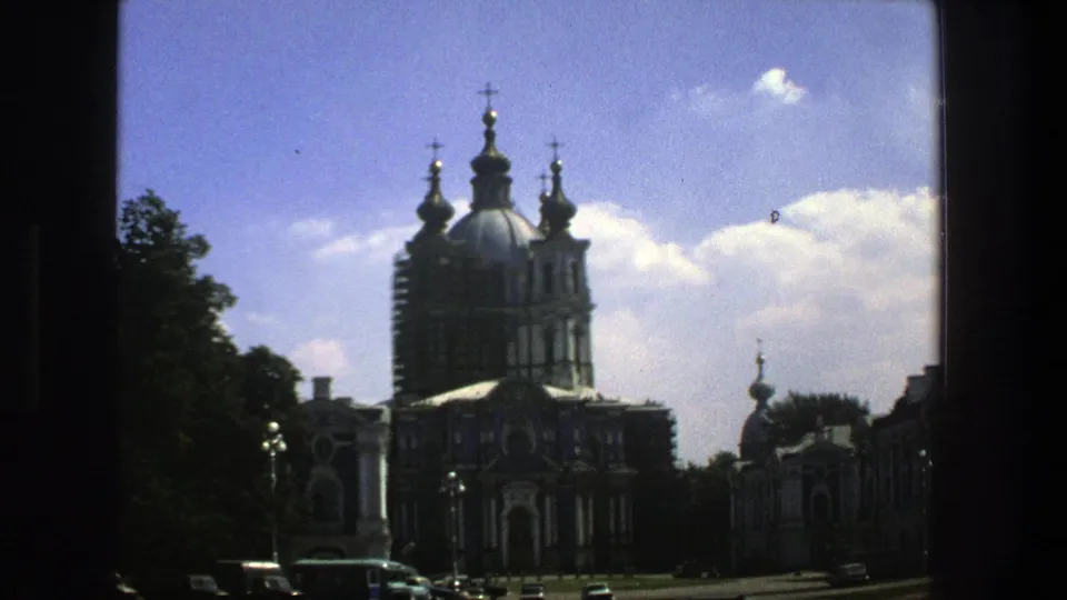 1975 8mm view in Denmark showing a large Orthodox church with blue onion domes, gold crosses, trees, parked cars, and blue sky.
