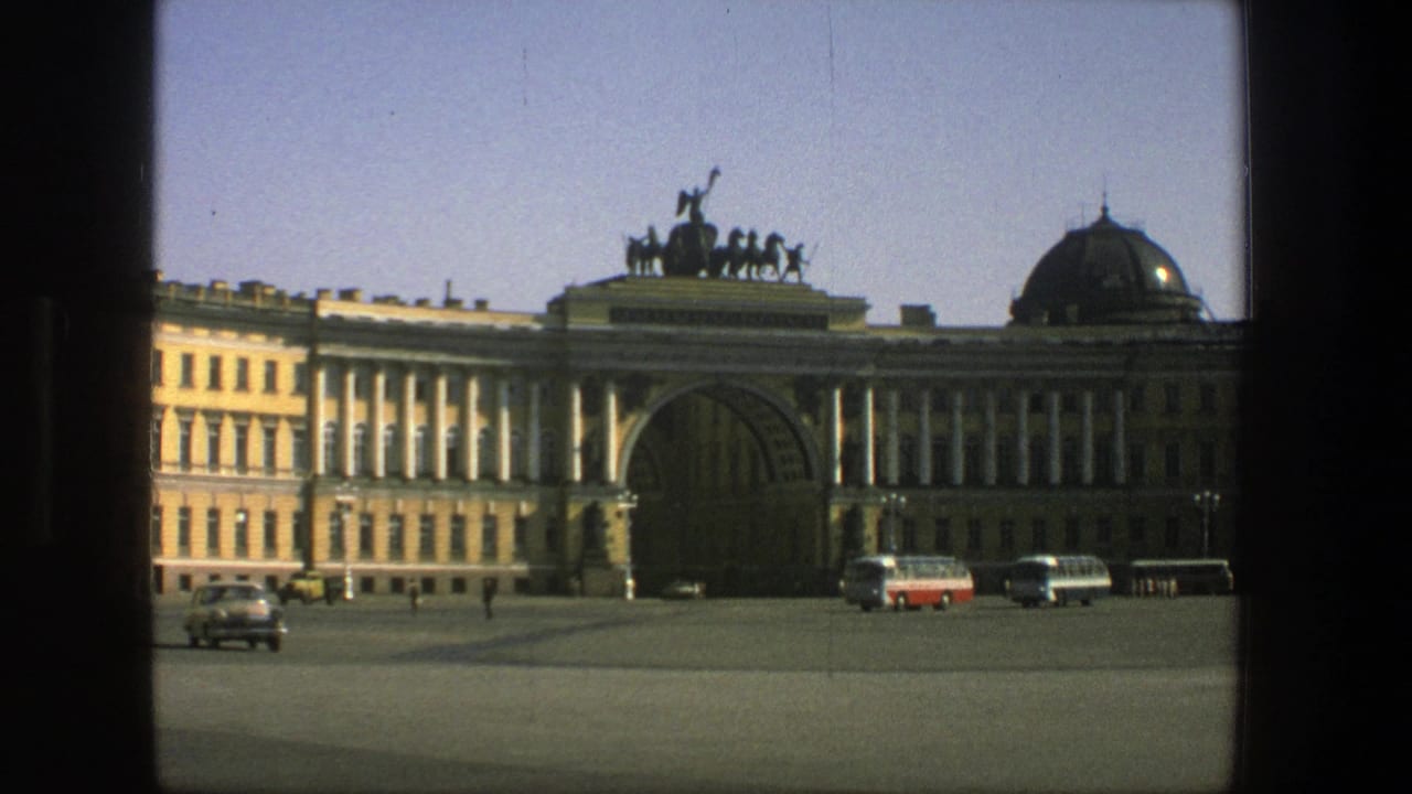1975 8mm view in Denmark of a broad plaza before a classical building, with parked buses, cars, and small pedestrians visible.