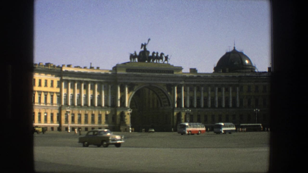 Light-colored sedan crosses the plaza in front of a classical building with buses parked near the arch.
